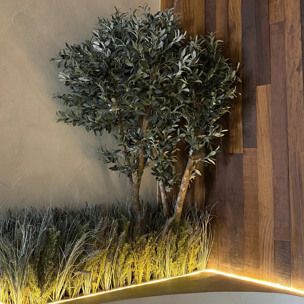 Olivo artificial hiperrealista sobre una pared de madera con gramíneas a los pies